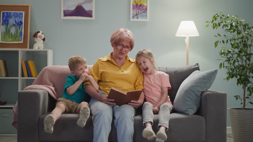 Cute old lady with glasses reads book to children at night, the children listen to fairy tales with interest, sit on gray sofa with blanket in cozy bright room, the child yawns and wants to sleep | Shutterstock HD Video #1098334113