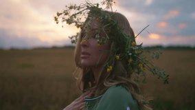 Beautiful woman with floral crown walks thoughtfully at wheat field. Feeling sad, cold and hopeless, wishing of peace. Bright make up, embroidered green dress. Sunset out of the city. High quality 4k