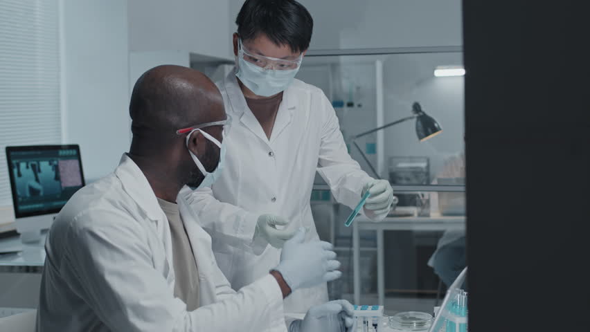 Medium side-view of Black male scientist in white gown and face mask using laptop computer, sitting at desk in modern laboratory, his female Asian colleague bringing him sample in tube | Shutterstock HD Video #1098343399