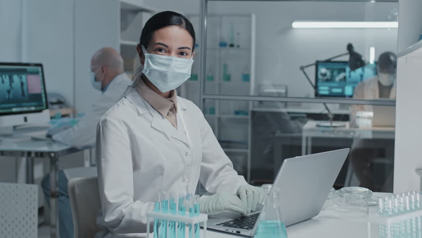 Waist-up portrait of young brown-eyed female lab technician in white gown, face mask and gloves sitting at desk with portable computer, looking on camera | Shutterstock HD Video #1098343963