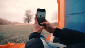 Young guy sits in tent in nature and shoots video on smart phone or takes photo of outdoors landscape with river or lake using camera. Male breaks camp on hike or picnic, sits under an awning alone.