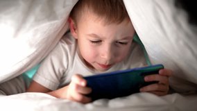 Bottom view of child who wrapped up in blanket and plays mobile phone. Online social networks. Smile and happy. Leisure activity in childhood. Holidays and rest. Baby boy watching and touch at screen.