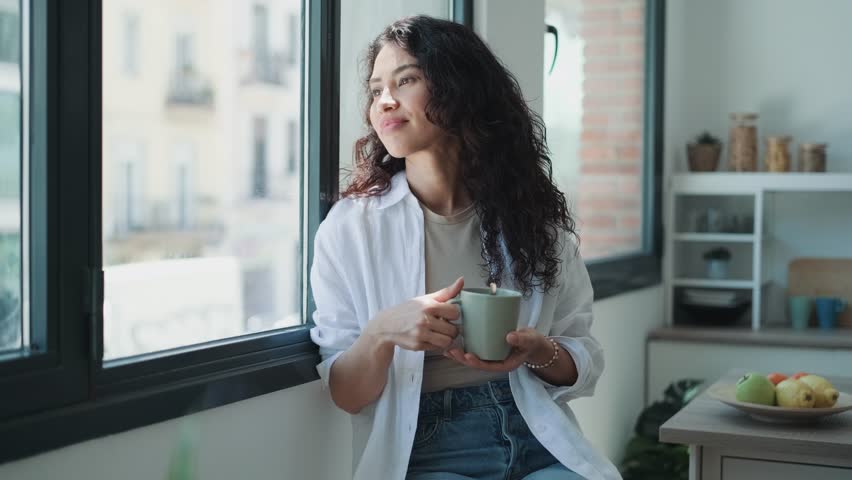 Video of beautiful woman drinking a cup of coffee while looking forward standing next to the window at home. | Shutterstock HD Video #1098349367