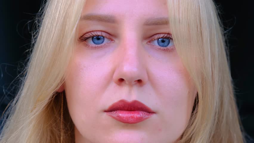 Portrait of a young beautiful sad blue-eyed blonde woman looking at camera against dark background. Hair flutters with the wind. Girl closes her eyes and experiences different experiences inside.