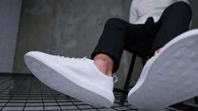Male sitting on the chair demonstrating modern sport footwear. Unrecognized man in white shirt and black trousers presents white sneakers in front of camera. Close up. Blurred backdrop.