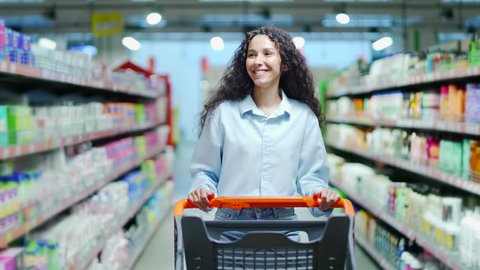 Happy young brunette woman with a shopping cart is shopping in a supermarket walking in hypermarket and pushing trolley going to buy buying and choosing household chemicals between the rows, videoclip de stoc