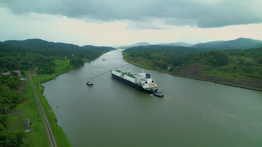 AERIAL: Flying along the shores of Panama Canal as a large cargo ship gets assisted along the wide river. Massive freight ship is assisted along the breathtaking Panama channel by a smaller ship. Royalty-Free Stock Footage #1098358663