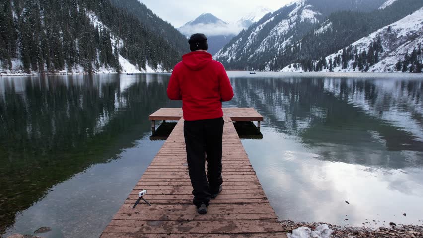 A guy walks along a pier on a mountain lake. Dark water like a mirror reflects snowy mountains, forest and clouds. Yellow sunset. A place for meditation and enjoyment. Epic. Lake Kolsai, Kazakhstan.