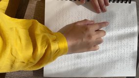 Vertical video. Top view. Woman reads text in Louis Braille book page. International Day of visual impairment and low vision. Female hand with yellow sleeve. January 4 is World Sight Day. Read. Finger