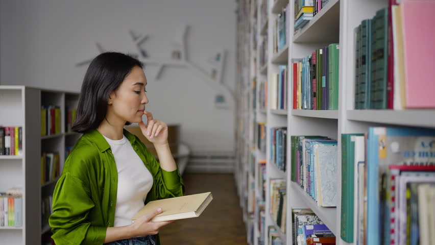 Young teacher picks up books from shelves in library thoughtfully. Asian woman looks for information to conduct interesting classes for students Royalty-Free Stock Footage #1098367437