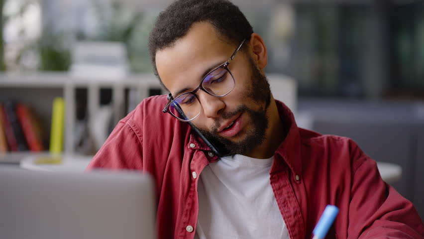 Close up portrait of pleasant positive African American university graduate student, business coach, prosperous teacher talking on mobile phone while working remotely on laptop during an online class