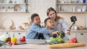 Excited african boys with caucasian mother having fun while making photos using selfie stick sitting at wooden table in kitchen, multiracial family having fun preparing organic salad from broccoli.
