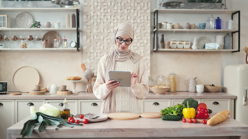 Happy muslim woman in hijab cooking healthy food pointing on vegetables lying on counter while reading recipe on digital tablet in modern kitchen at home. Preparing vegetable salad.