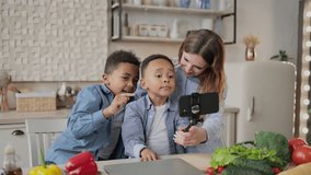 Excited african boys with caucasian mother having fun while making photos using selfie stick sitting at wooden table in kitchen, multiracial family having fun preparing organic salad.