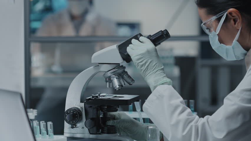 Side view of young female scientist in protective goggles, gloves and mask using microscope in modern laboratory | Shutterstock HD Video #1098370669