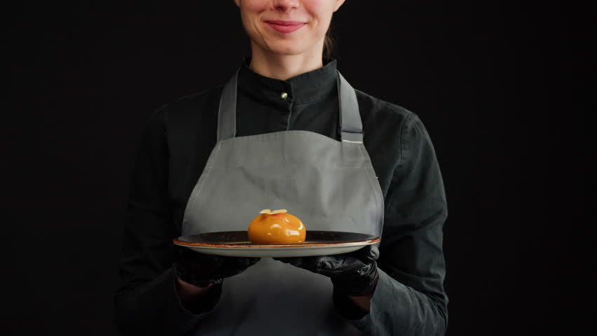 Woman chef holding mousse orange cake, creamy dessert close-up. Almond dacquoise, raspberry confit, crispy layer with caramelized hazelnuts and raspberry powder, mousse based on bitter chocolate. Royalty-Free Stock Footage #1098375153