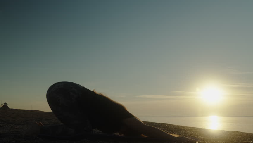 Yoga at dawn by the sea. The girl bends in the pose of a dog. Silhouette opposite the sun.