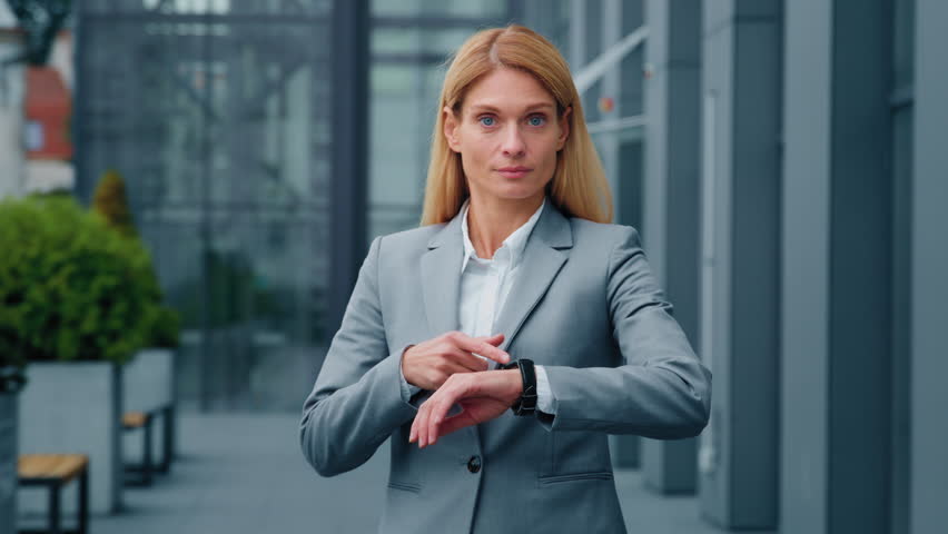 Serious punctual confident businesswoman looking at wristwatch checking hour point to time young caucasian woman standing outdoors near office building waiting for meeting dissatisfied with being late | Shutterstock HD Video #1098379427