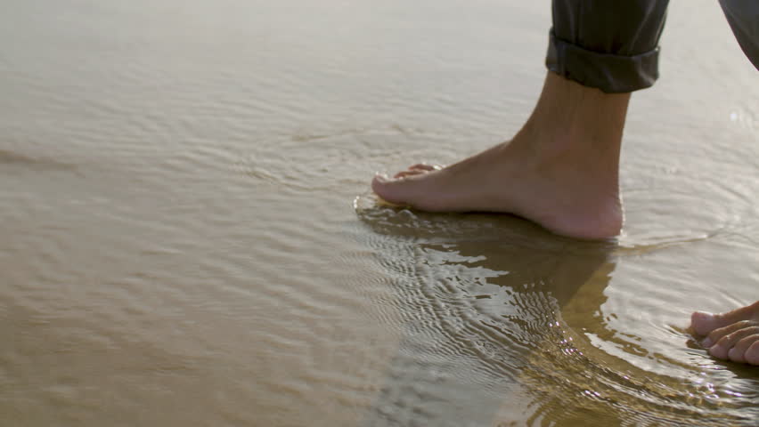 Legs closeup of Caucasian man walking alone seashore, stepping on wet sand. Side view, slow motion shot. Leisure activity, holiday concept.