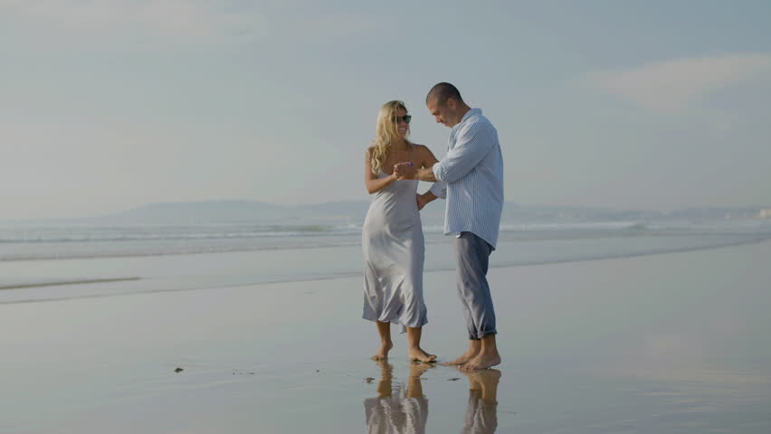 Beautiful young couple dancing at seashore, spending time together on holiday at sunset. Caucasian man in blue shirt twirling attractive blonde woman in sunglasses under his arm. Holiday, love concept