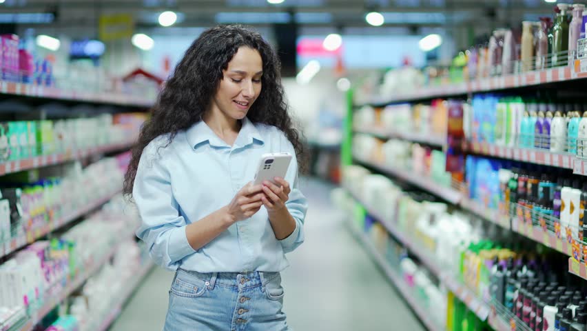 Happy young hispanic woman uses mobile phone in a supermarket while standing between the rows. Indoor Female brunette typing text message, chatting browsing in smartphone in grocery store food market Royalty-Free Stock Footage #1098389727