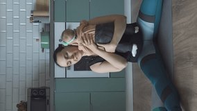 Vertical video. Young mother working out while lying on exercising mat with cute baby in the kitchen at home