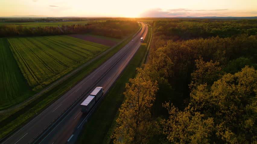AERIAL: Convoy of cargo trucks on a countryside highway in beautiful golden light. Low traffic on a motorway that crosses beautiful landscape in gorgeous light. Truck transport for delivering goods.