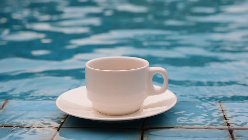 A white cup of coffee lays on the edge of the swimming pool.