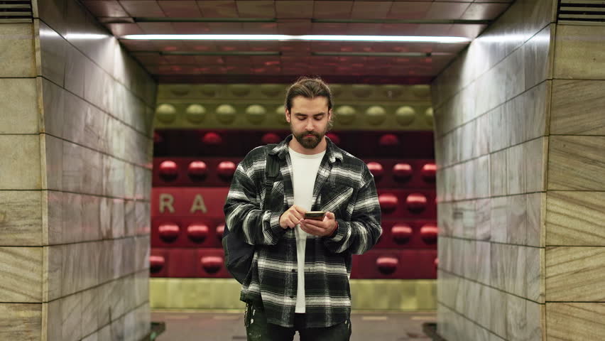 Front view of handsome tourist traveling by public transport, standing in subway, waiting. Brunette man with beard holding, using smartphone, reading. Concept of traveling.