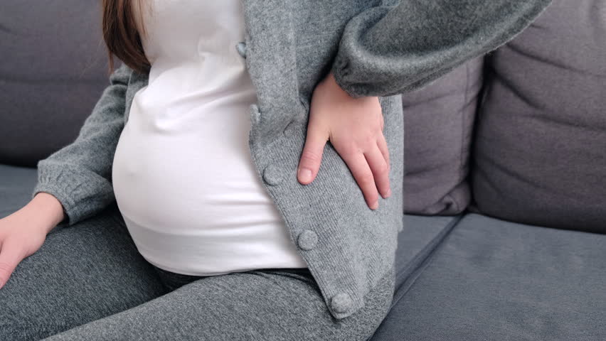 Close up side view of young pregnant woman massaging back muscles sitting on sofa, suffering from severe painful feelings, female 20s  with third trimester big belly having joints strain in lower back | Shutterstock HD Video #1098410757