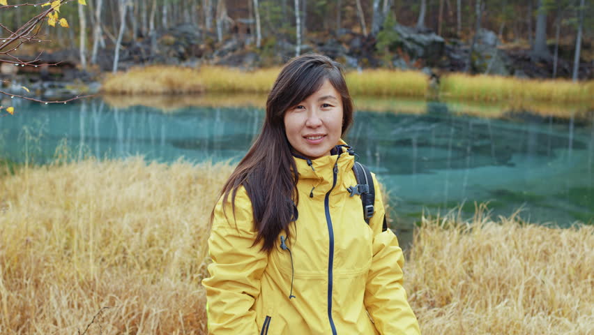 Beautiful girl stands in front of geyser lake surrounded by yellowed reeds. Young woman with oriental appearance is near blue pond. Portrait of long-haired brunette looking to camera in forest.