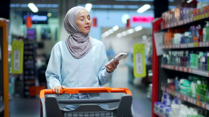 Young muslim woman in hijab Buyer customer checking shopping list using smartphone, purchasing in grocery store in supermarket female shopper pick use mobile phone in hypermarket or food market Royalty-Free Stock Footage #1098412571