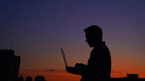 Man IT engineer or freelancer in silhouette working outdoor using laptop. Sunset time. Adorable dusk at background. Hardly working evening. High quality 4k video footage