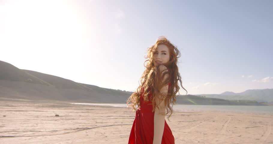 Gorgeous caucasian girl wearing a red dress is posing for a photoshoot. Attractive woman gender Z on scenic Surreal landscape background while wind is blowing her red hair. Ginger Lady enjoying moment