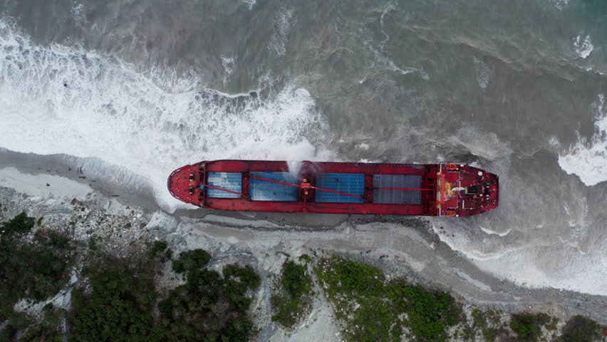 Big red cargo ship ran aground. Freighter went ashore. Bulk carrier got upon bank. Watercraft is on seashore near mountains. Barge was stranded. Navigation, shipping. Top view