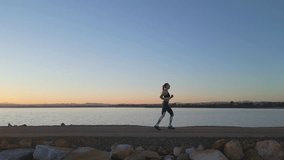  Fit young female athlete running jogging on the pier in a calm water with a lake and beautiful sunset. Drone footage. Slow motion. Sport workout training motivation video. Side view