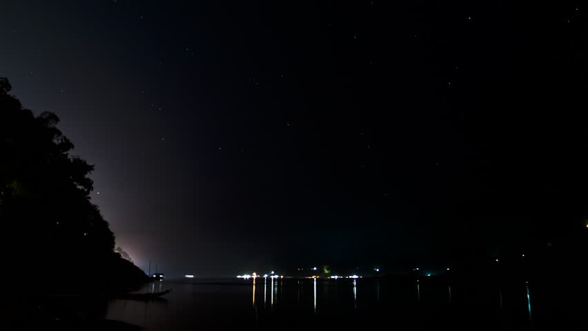 Timelapse the stars and small village lights are visible, and a person starts a boat and motors away in the foreground. 