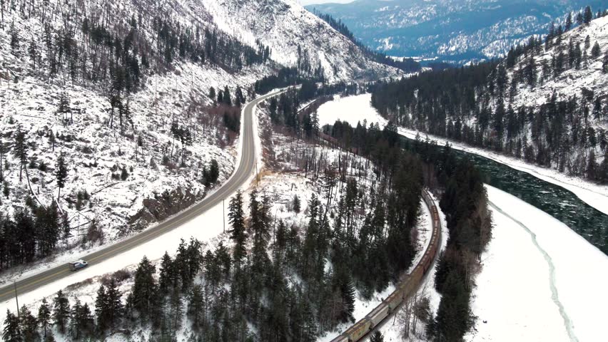 A Medium Aerial Shot of Cars and Freight Train on Yellowhead Highway 5 and Railway Through the Frozen North Thompson River Valley near Kamloops, Magnificent Mountainous Snow-covered Landscape Royalty-Free Stock Footage #1098437277