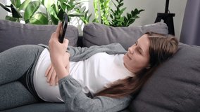 Relaxed peaceful smiling young woman with big belly lying on comfy sofa and using modern smartphone. Pregnant lady laughing while watching some funny videos. Leisure activity of future mother concept