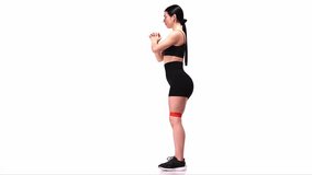 Side view. Young sportive girl training with fitness elastic bands over white studio background. Doing squats. Modern sport, action, motion, youth concept. Fitness, hobby, healthy lifestyle