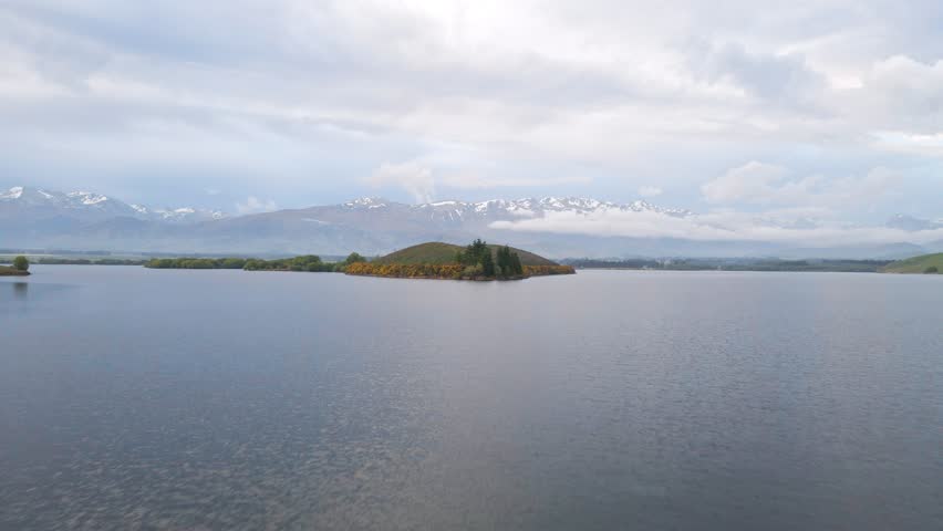 Island on a lake with southern New Zealand alps in background. Fast, low aerial approach