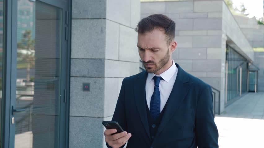 Sad entrepreneur reading bad news on mobile phone walking downtown. Worried bearded man entrepreneur in formal suit looking on mobile phone feeling sad from negative news outdoors. Overworking concept | Shutterstock HD Video #1098455097