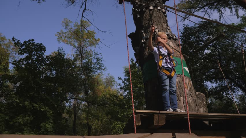 A little boy in climbing gear is walking along a rope road in an adventure Park, holding on to a rope and a carabiner.