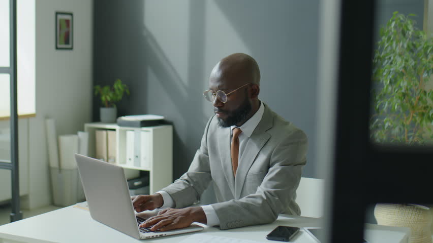Medium shot of African American businessman in formalwear and eyeglasses using laptop while working at desk in modern office | Shutterstock HD Video #1098457619