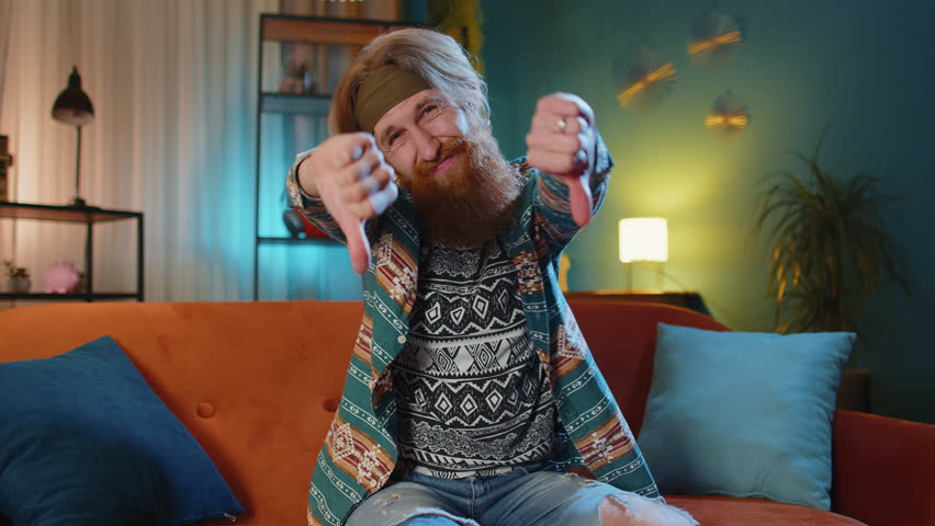 Dislike. Upset man showing thumbs down sign gesture, expressing discontent, disapproval, dissatisfied bad work at modern home apartment indoors. Displeased hippie guy in living room sitting on sofa | Shutterstock HD Video #1098457773
