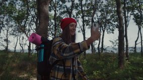 Close-up of woman solo traveller on hike in epic forest. Happy and excited young woman in with black touring backpack on her back walks through the woods overlooking the seascape and talks