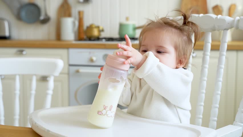 Cute messy baby girl crying while drinks powered milk from a bottle at home kitchen. Children liquid food. Concept of artificial or breastmilk nourishment. | Shutterstock HD Video #1098460889