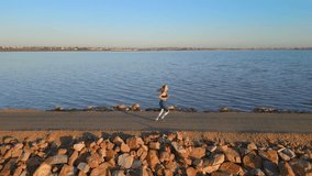 Fit young female athlete running on the pier in a calm water of a salt lake and beautiful sunset. Drone footage. Slow motion. Sport workout training motivation video. Side view