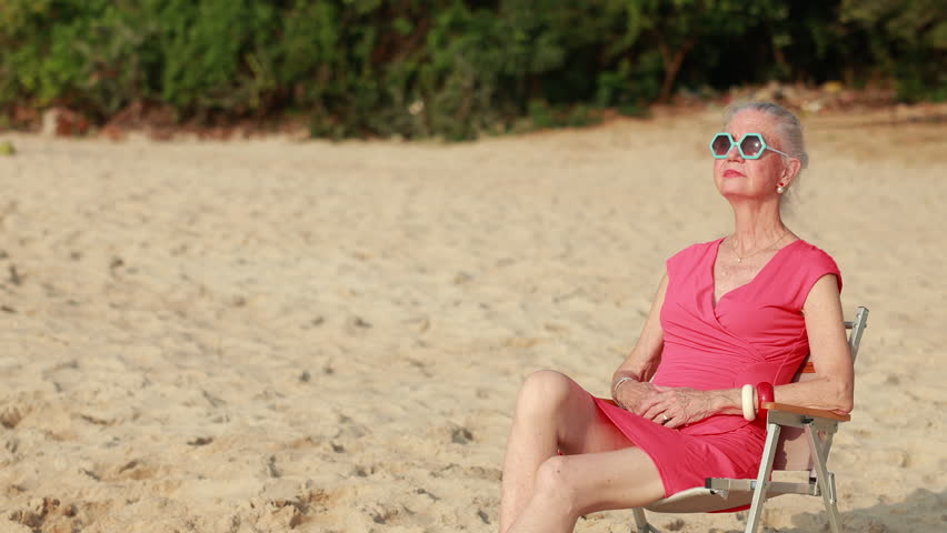 Elderly woman wearing fashion sunglasses looking at the sea sky sitting on chair on beach. Vacation trip summer holiday.