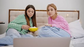 Close-up two beautiful teens girls, sharing happy moments together, eating popcorn and watching comedy on laptop, lying on the bed in a cozy homely friendly atmosphere. Adolescence Youth Friendships